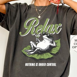 May include: Pepper-colored t-shirt with a graphic of a frog relaxing on a lily pad, holding a cocktail. The word "Relax" is arched above the frog, and the phrase "Nothing is Under Control" is below. "More color options" is also displayed.