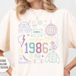 May include: Ivory-colored t-shirt with a colorful 1986 graphic. The design includes a boombox, cassette tape, disco ball, and other vintage elements. The shirt has a relaxed fit and short sleeves.