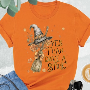 May include: An orange short-sleeved t-shirt featuring a graphic of a grey witch hat and a broomstick with autumn leaves. The text reads, "YES I CAN DRIVE A STICK." The shirt is displayed with blue jeans, a black crossbody bag, and black sneakers.