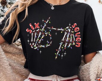 May include: Black t-shirt with a skeleton hand design holding Christmas lights and the text "Six Seven". The person is wearing red and white plaid pajama pants and holding a mug with a Santa Claus design.