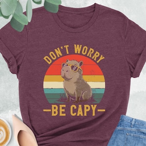 May include: Heather maroon t-shirt with a graphic of a capybara wearing sunglasses, set against a sunset design. The text reads "DON'T WORRY - BE CAPY". The shirt is paired with blue jeans and nude-colored shoes.