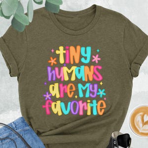 May include: Heather olive green t-shirt with the text "TINY HUMANS are my favorite" in colorful letters. The shirt is styled with blue jeans, a black crossbody bag, and black high-top sneakers. A cup of coffee with latte art is also present.
