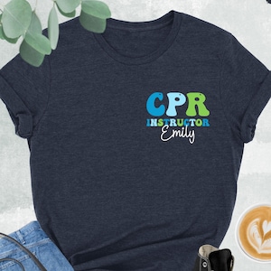 May include: A heather navy blue t-shirt with the text "CPR Instructor Emily" in colorful, retro-style letters.