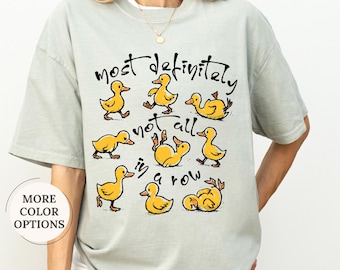 May include: A light green t-shirt with the phrase "most definitely not all in a row" and illustrations of yellow ducklings. The shirt has a relaxed fit and short sleeves.