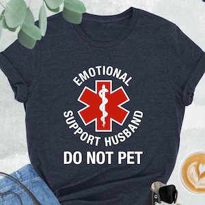 May include: Heather navy t-shirt with the text "EMOTIONAL SUPPORT HUSBAND DO NOT PET" in white. The design includes a red and white medical symbol. The shirt is paired with blue jeans, a black purse, and black high-top sneakers.