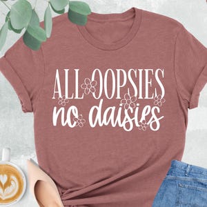 May include: A heather mauve t-shirt with the text "ALL OOPSIES no daisies" in white lettering. The text is surrounded by small white flower graphics.