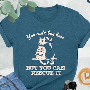 May include: A teal blue t-shirt with the text "You can't buy love but you can rescue it" and a graphic of two cats hugging.