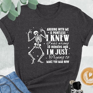 May include: Dark grey heather t-shirt with a skeleton graphic and the text "ARGUING WITH ME IS POINTLESS I KNEW I was wrong 10 minutes ago I'M JUST Trying to MAKE YOU MAD NOW". The shirt is paired with blue jeans, a black purse, and black high-top sneakers.
