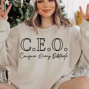 May include: A beige sweatshirt with the text "C.E.O. Conquer Every Obstacle" in black lettering.