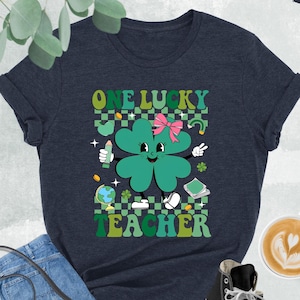 May include: A navy blue t-shirt with a green four-leaf clover cartoon character wearing a pink bow. The clover is holding a pencil and a globe. The text "ONE LUCKY TEACHER" is printed in green and yellow letters.
