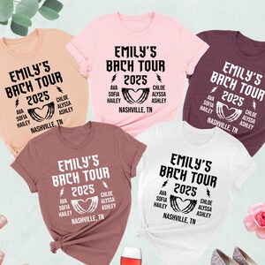 May include: Five t-shirts in various colors, including pink, white, and maroon, are displayed. Each shirt features the text "EMILY'S BACH TOUR 2025" with a heart design and the names of the tour members. The shirts are arranged with a rose, wine, and shoes.