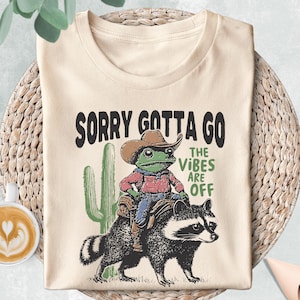 May include: Cream-colored t-shirt with a graphic featuring a cowboy frog riding a raccoon, with a cactus and the text "SORRY GOTTA GO THE VIBES ARE OFF". The shirt is laid on a woven mat.