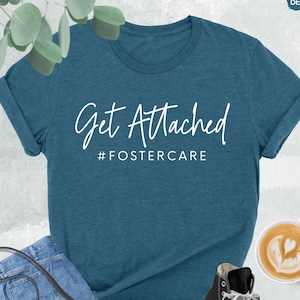 May include: A teal blue t-shirt with the text "Get Attached #FOSTERCARE" in white lettering. The shirt is photographed on a white background with jeans, a purse, and a cup of coffee.