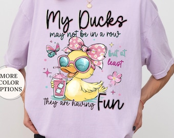 May include: Orchid-colored t-shirt with a graphic of a cartoon duck wearing sunglasses and a bow, holding a drink. Text reads "My Ducks may not be in a row but at least they are having Fun."