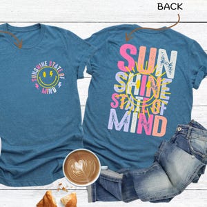 Pu&ograve; includere: T-shirt color verde acqua con la scritta "Sunshine State of Mind" sul davanti e sul retro. Il davanti presenta una faccina sorridente e il retro le parole in un design colorato. Scarpe da ginnastica bianche e jeans accanto alle magliette.
