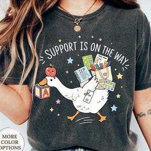 May include: A charcoal gray t-shirt featuring a graphic of a white goose carrying a tote bag filled with school supplies, an apple, and a block with the letter A. The text "SUPPORT IS ON THE WAY" is printed above the goose, surrounded by colorful stars.