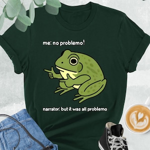 May include: A dark green t-shirt with a cartoon frog illustration and the text "me: no problemo!" and "narrator: but it was all problemo". The shirt is paired with blue jeans, a black purse, and black high-top sneakers.