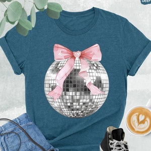 May include: A teal blue t-shirt with a silver disco ball and a pink bow on it. The shirt is laying on a white surface with a pair of black sneakers, a denim purse, and a cup of coffee.