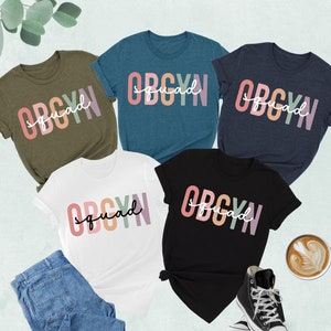 May include: Five t-shirts in different colors with the text "OBGYN squad" printed in rainbow colors. The shirts are laid out on a white surface with a cup of coffee and a pair of sneakers.