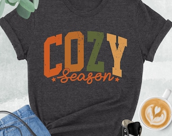 May include: Dark grey heather t-shirt with the words "COZY Season" in orange, green, and yellow. The shirt is paired with blue jeans, a black purse, and black high-top sneakers. A cup of coffee with latte art is also in the image.