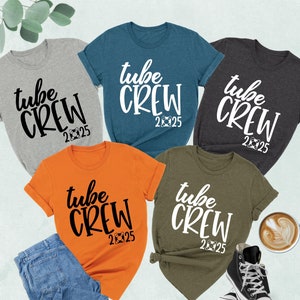 May include: Five t-shirts in different colors with the text "tube crew 2025" printed on them. The shirts are laid out on a surface with a cup of coffee and a pair of sneakers.