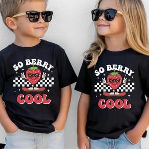 May include: Two children wearing black t-shirts with a strawberry graphic. The shirts have the text "SO BERRY COOL" in white and red. The strawberry is wearing sunglasses and making a peace sign. The children are also wearing sunglasses.