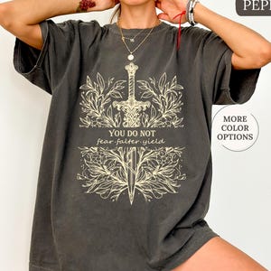 May include: A dark gray oversized t-shirt with a white graphic of a sword surrounded by floral designs. The text "You do not fear, falter, yield" is printed on the shirt.