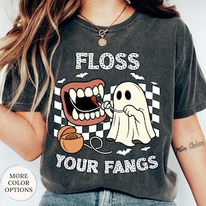May include: A charcoal gray t-shirt featuring a graphic print of a white ghost flossing its fangs with dental floss. The design includes a black and white checkered background, small bats, and the text FLOSS YOUR FANGS in a white spotted font.