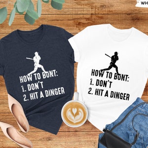 May include: Two t-shirts with a baseball player graphic and the text "How to Bunt: 1. Don't 2. Hit a Dinger". The shirts are in heather navy and white.