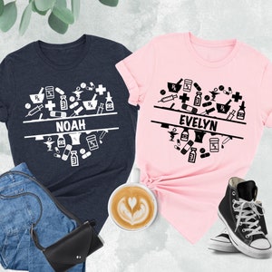 Puede incluir: Dos camisetas con un diseño de corazón hecho de símbolos médicos y los nombres Noah y Evelyn. Las camisetas son de color azul marino jaspeado y rosa.