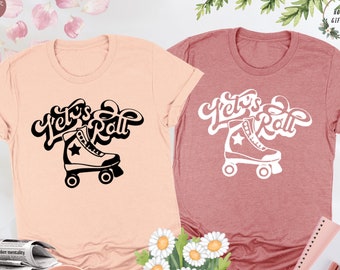 May include: Two t-shirts with the text "Let's Roll" in a retro font, with a roller skate graphic. The t-shirt on the left is peach colored and the t-shirt on the right is a dusty rose color.
