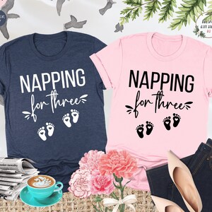 Może przedstawiać: Dwie koszulki, jedna różowa, a druga granatowa, z napisem "NAPPING for three" i trzema śladami stóp z sercami na każdym. Koszulki leżą na stole z bukietem różowych kwiatów, filiżanką kawy i gazetą.