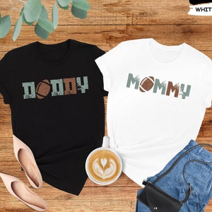 May include: Two black and white t-shirts with the words "Daddy" and "Mommy" printed on them. Each shirt has a football graphic.