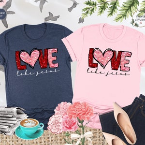 May include: Two t-shirts with the words "Love like Jesus" in red and pink letters with a heart shape in the middle. The heart is made of pink and red sequins. The shirts are on a table with a bouquet of pink flowers and a cup of coffee.