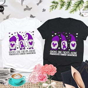 May include: Two t-shirts with a design of three gnomes holding purple hearts. The gnomes are wearing purple hats and have purple beards. The text on the shirts reads "Gnome One Fights Alone Pancreatic Cancer Awareness".