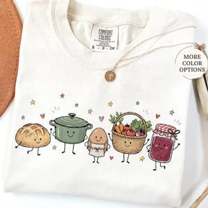 May include: An ivory t-shirt featuring a graphic print of five anthropomorphic food items: a loaf of bread, a green pot, an egg wearing an apron, a basket of vegetables, and a jar of jam. The shirt has a Comfort Colors label at the neck.