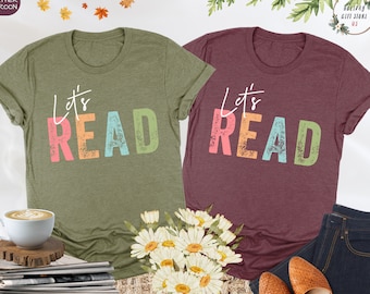May include: Two t-shirts with the text "Let's Read" in colourful, distressed lettering. The shirts are in heather olive and heather maroon.