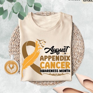 May include: A beige t-shirt with a brown ribbon and a yellow feather design. The text "August is Appendix Cancer Awareness Month" is printed on the shirt.