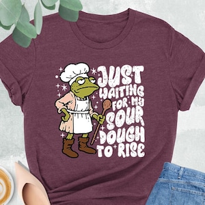 May include: Heather maroon t-shirt with a frog chef graphic. The frog wears a chef's hat, apron, and holds a wooden spoon. Text on the shirt reads "JUST WAITING FOR MY SOUR DOUGH TO RISE". The shirt is paired with jeans and shoes.