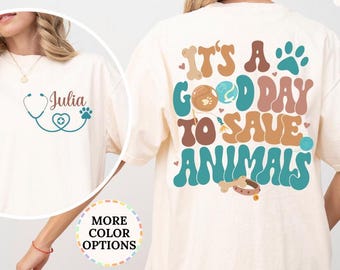 May include: Ivory-colored t-shirt with the text "IT'S A GOOD DAY TO SAVE ANIMALS" in a retro font. The design includes a paw print, a bone, and a stethoscope. The front of the shirt has a stethoscope design and the name "Julia".