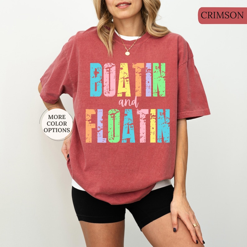 May include: Crimson t-shirt featuring the words "BOATIN and FLOATIN" in distressed, multi-coloured lettering. The shirt has a loose fit and is worn with black shorts. A button on the left says "MORE COLOR OPTIONS".