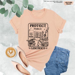 May include: A peach colored t-shirt with a black graphic design of a stack of books, a quill pen, and the text "Protect Public Libraries".