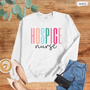 K&ouml;nnte beinhalten: Wei&szlig;er Pullover mit dem Text "Hospice nurse" in Regenbogenfarben.