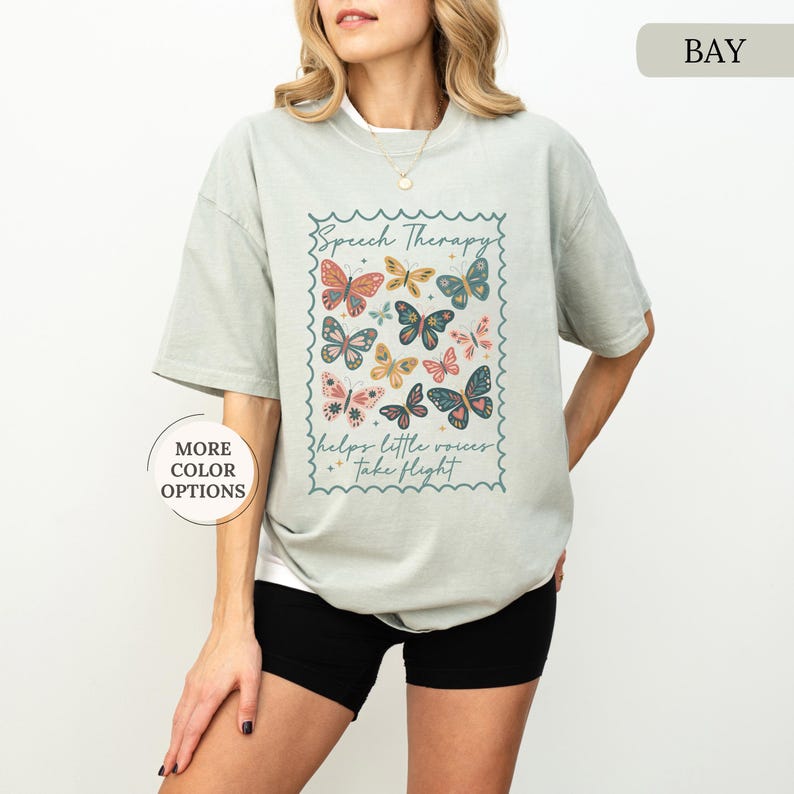 Puede incluir: Camiseta verde claro con un dise&ntilde;o de mariposas. El dise&ntilde;o incluye las palabras "Speech Therapy" y "helps little voices take flight". Las mariposas son de varios colores, incluyendo naranja, azul y rosa.