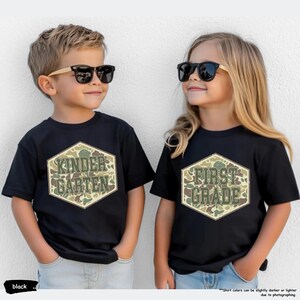 May include: Two children wearing black t-shirts and sunglasses. The shirts feature a camouflage hexagon design with the words "KINDERGARTEN" and "FIRST GRADE". The t-shirts are black.