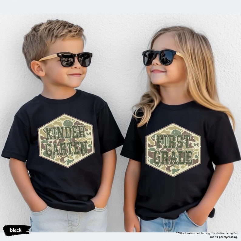 May include: Two children wearing black t-shirts and sunglasses. The shirts feature a camouflage hexagon design with the words "KINDERGARTEN" and "FIRST GRADE". The t-shirts are black.