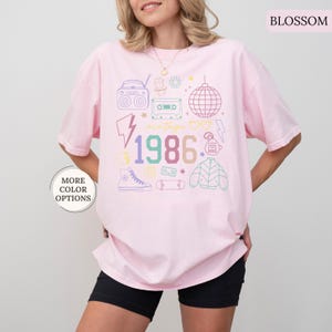 May include: Light pink t-shirt with a colorful vintage 1986 graphic. The design includes a boombox, cassette tape, disco ball, and other retro icons. The shirt has a relaxed fit and is made of a soft material.