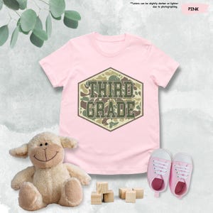 May include: A pink t-shirt featuring the text "THIRD GRADE" in a camouflage pattern. The image includes a stuffed lamb, wooden blocks, and pink baby shoes. The text "colors can be slightly darker or lighter due to photographing" is also visible.