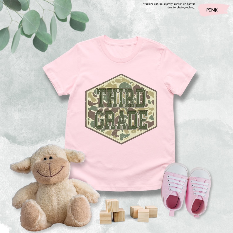 May include: A pink t-shirt featuring the text "THIRD GRADE" in a camouflage pattern. The image includes a stuffed lamb, wooden blocks, and pink baby shoes. The text "colors can be slightly darker or lighter due to photographing" is also visible.