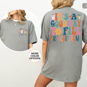 May include: A grey t-shirt featuring the phrase "IT'S A GOOD DAY TO FILL PRESCRIPTIONS" in a colourful, retro font on the back. The front displays a smaller design with a prescription pad, coffee cup, and the name "Sophia".